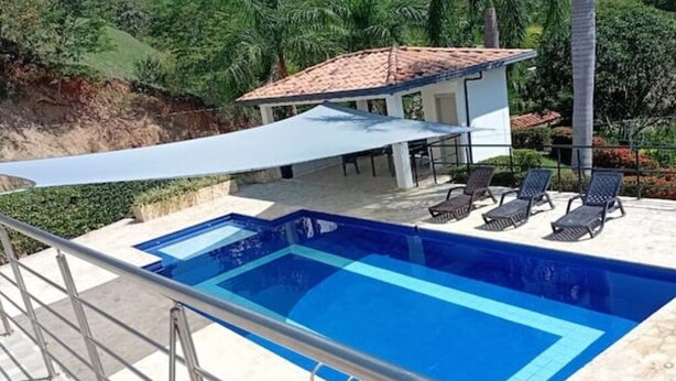 Una piscina con un toldo, sillas de descanso y un pequeño edificio con techo de tejas.