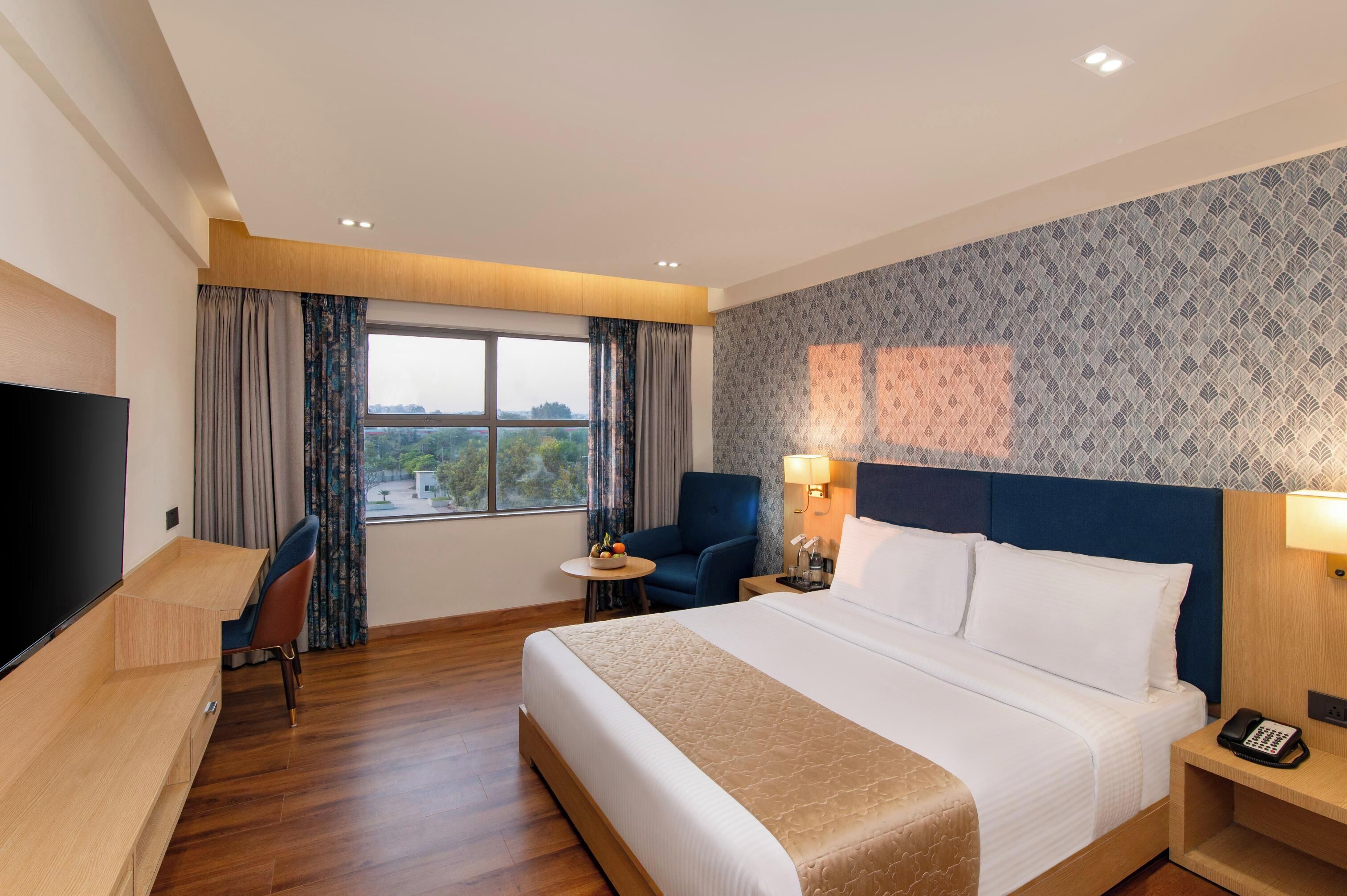 A hotel room with a bed, a desk, a chair, a small table, a TV, and a window with curtains.