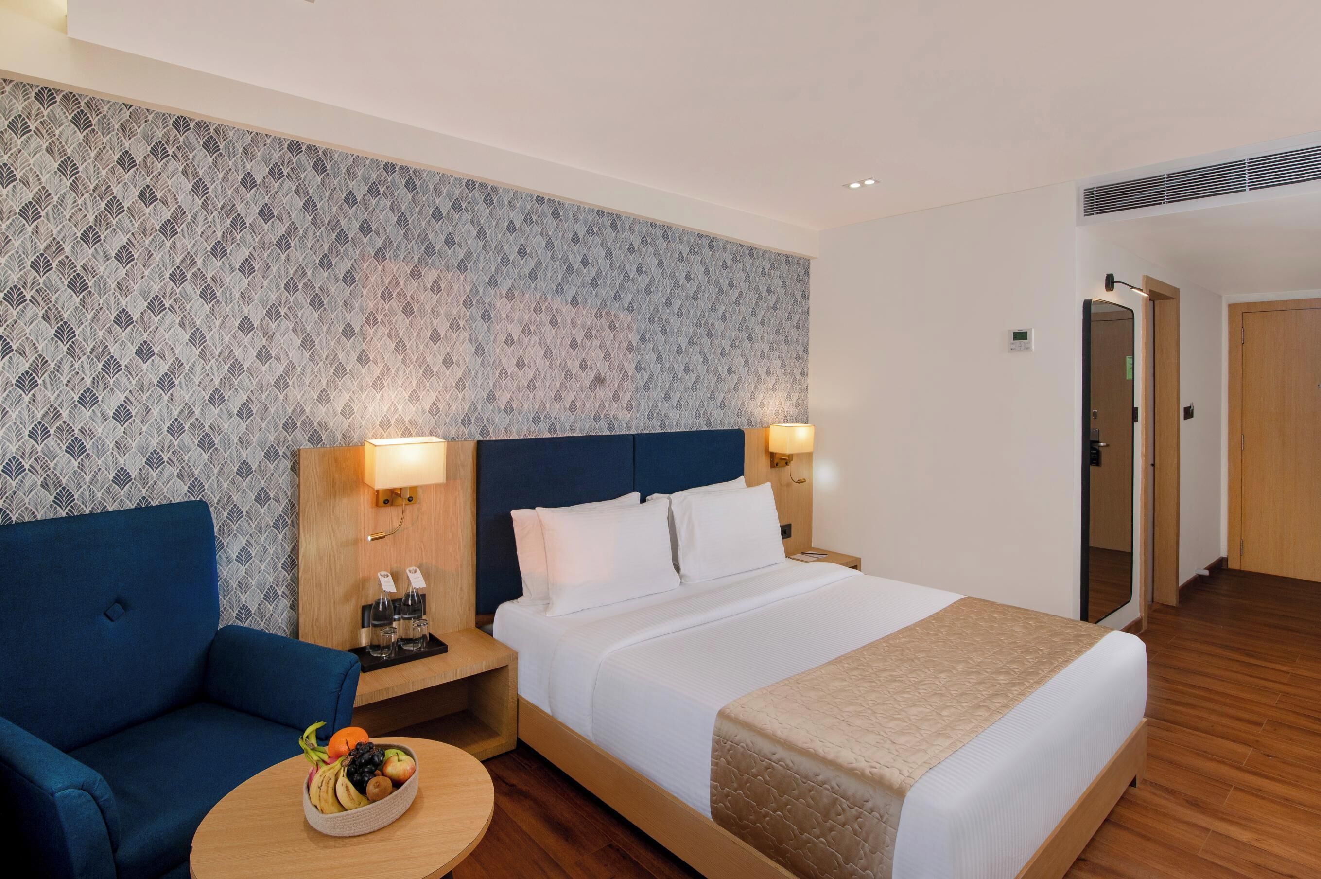 A hotel room with a bed, a blue sofa, a bedside table with a lamp, and a fruit bowl.