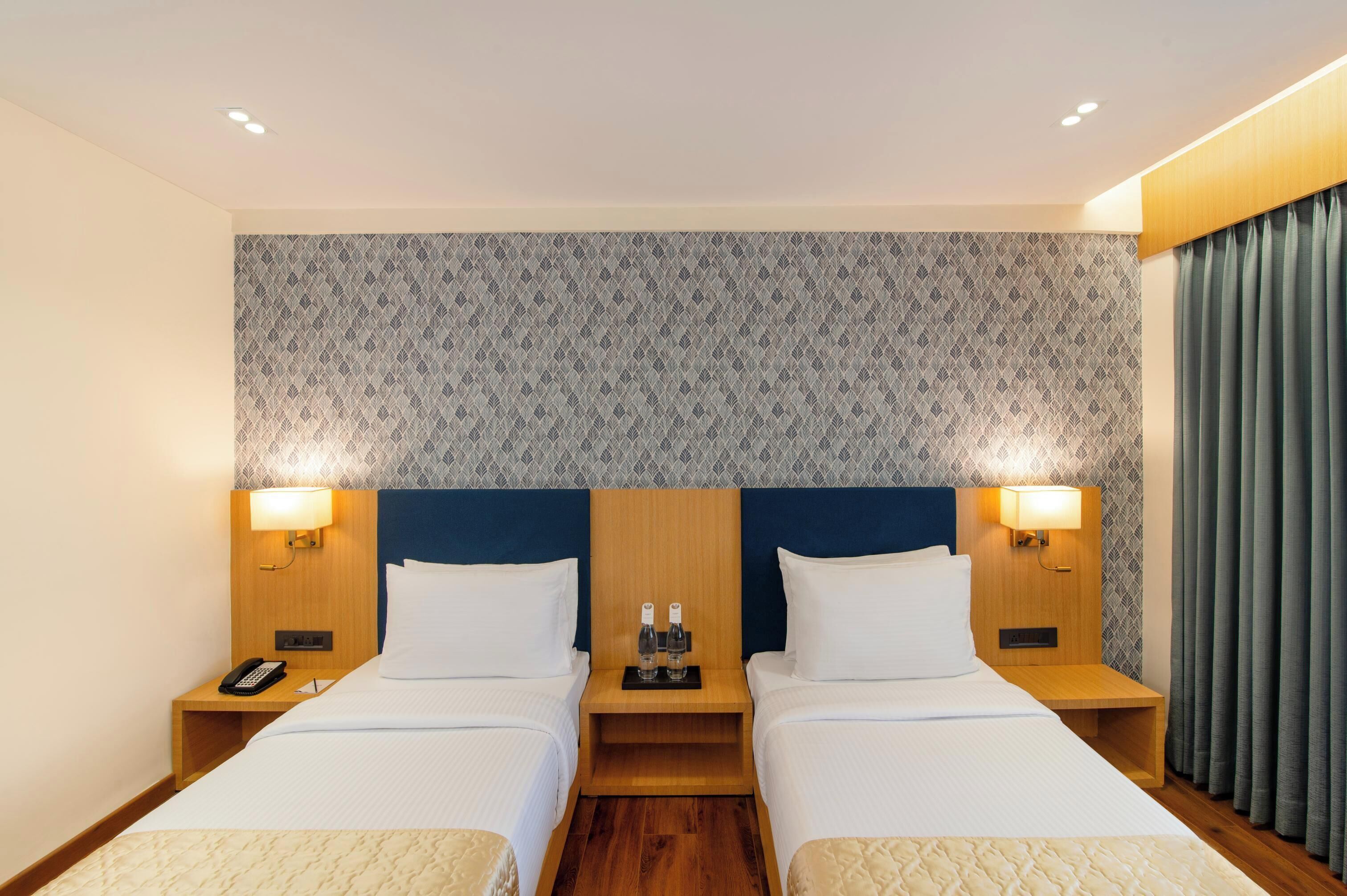 A hotel room with two beds, a patterned wall, wooden headboards, bedside lamps, a phone, and a nightstand with bottles.