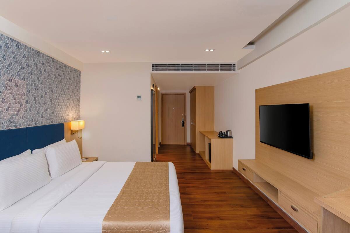 A modern hotel room with a large bed, a TV mounted on the wall, a desk with a chair, and a patterned wall.