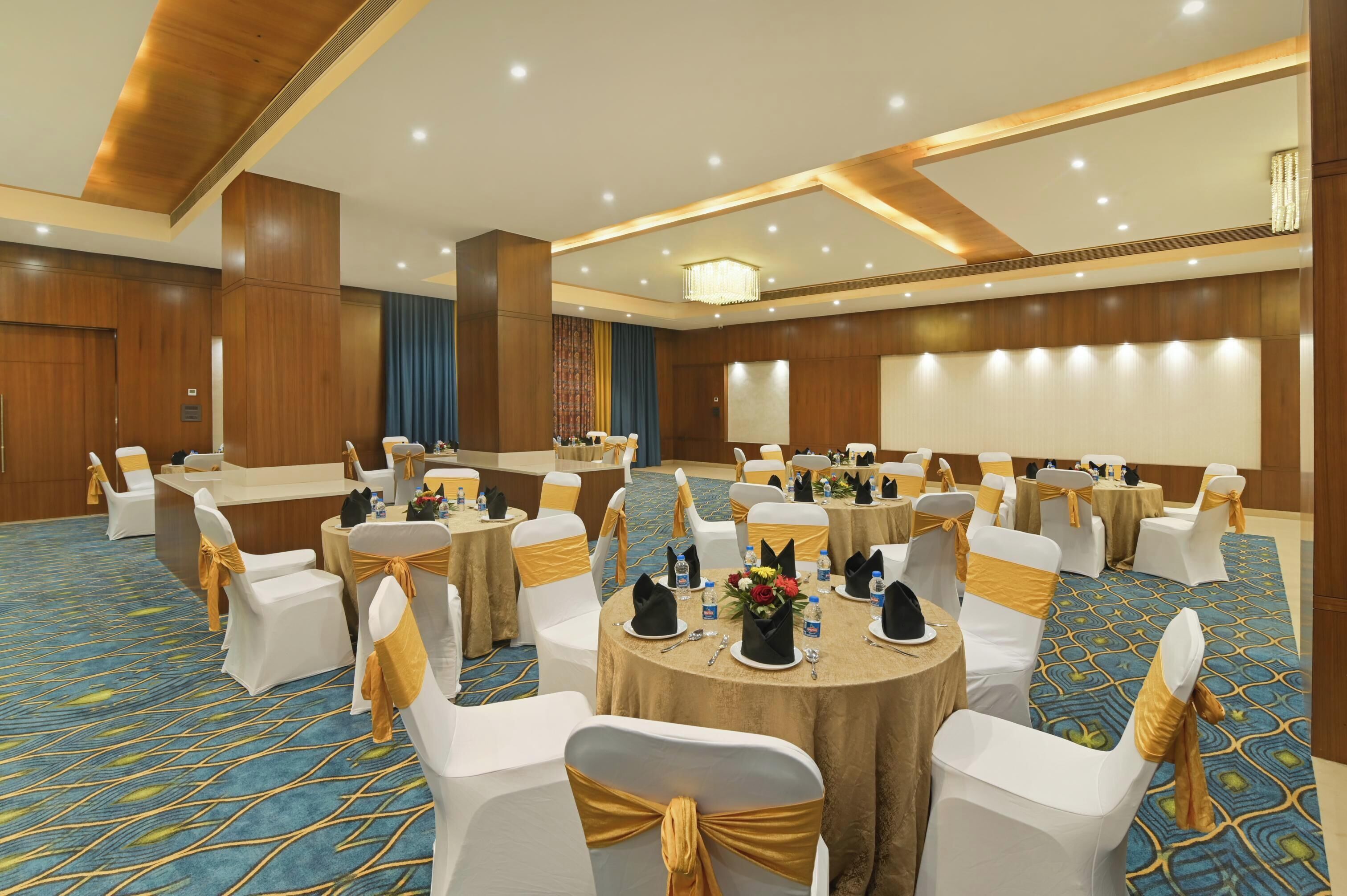 A banquet hall with round tables, white chairs with gold sashes, and a blue patterned carpet.
