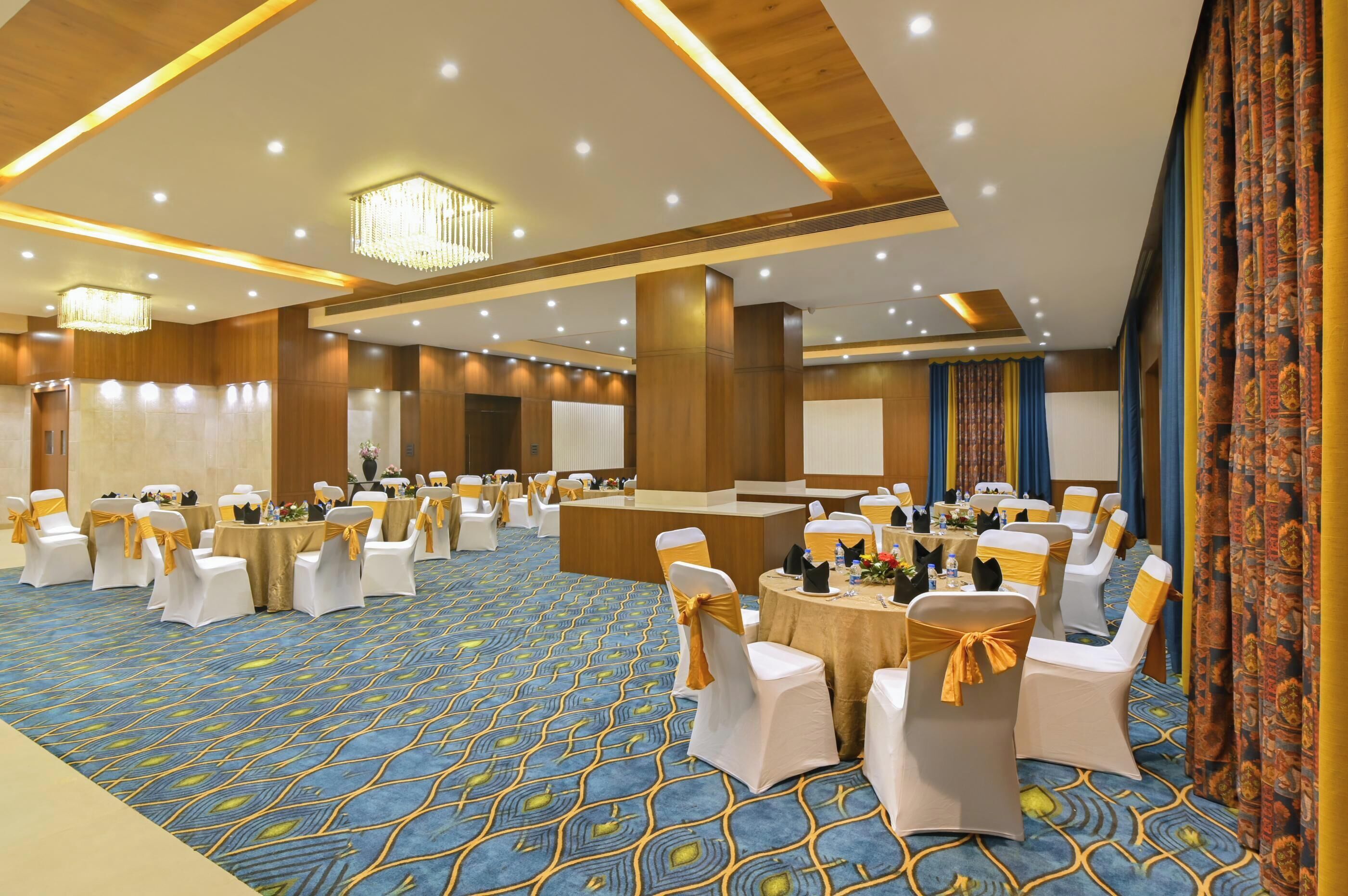 A banquet hall with round tables, white chairs with yellow sashes, and a blue patterned carpet.