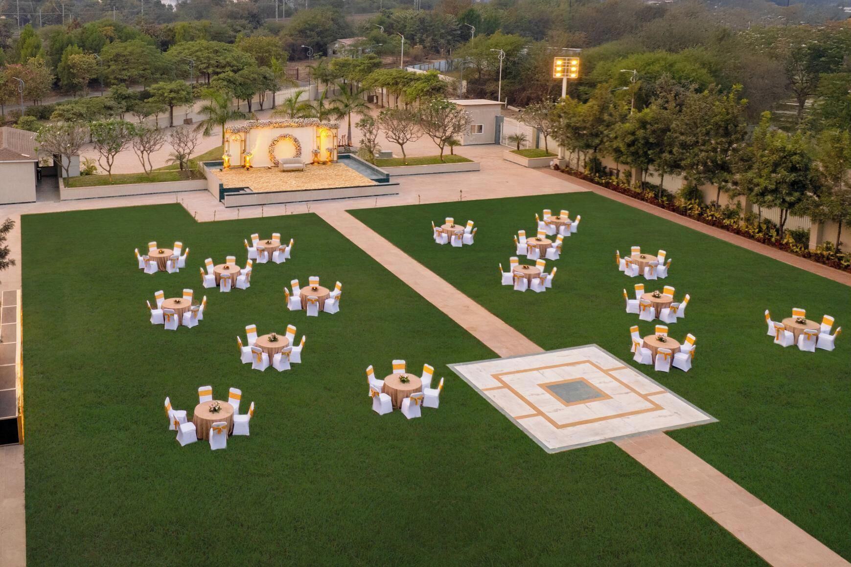 An outdoor event space with multiple round tables and chairs, a stage, and a well-maintained lawn.