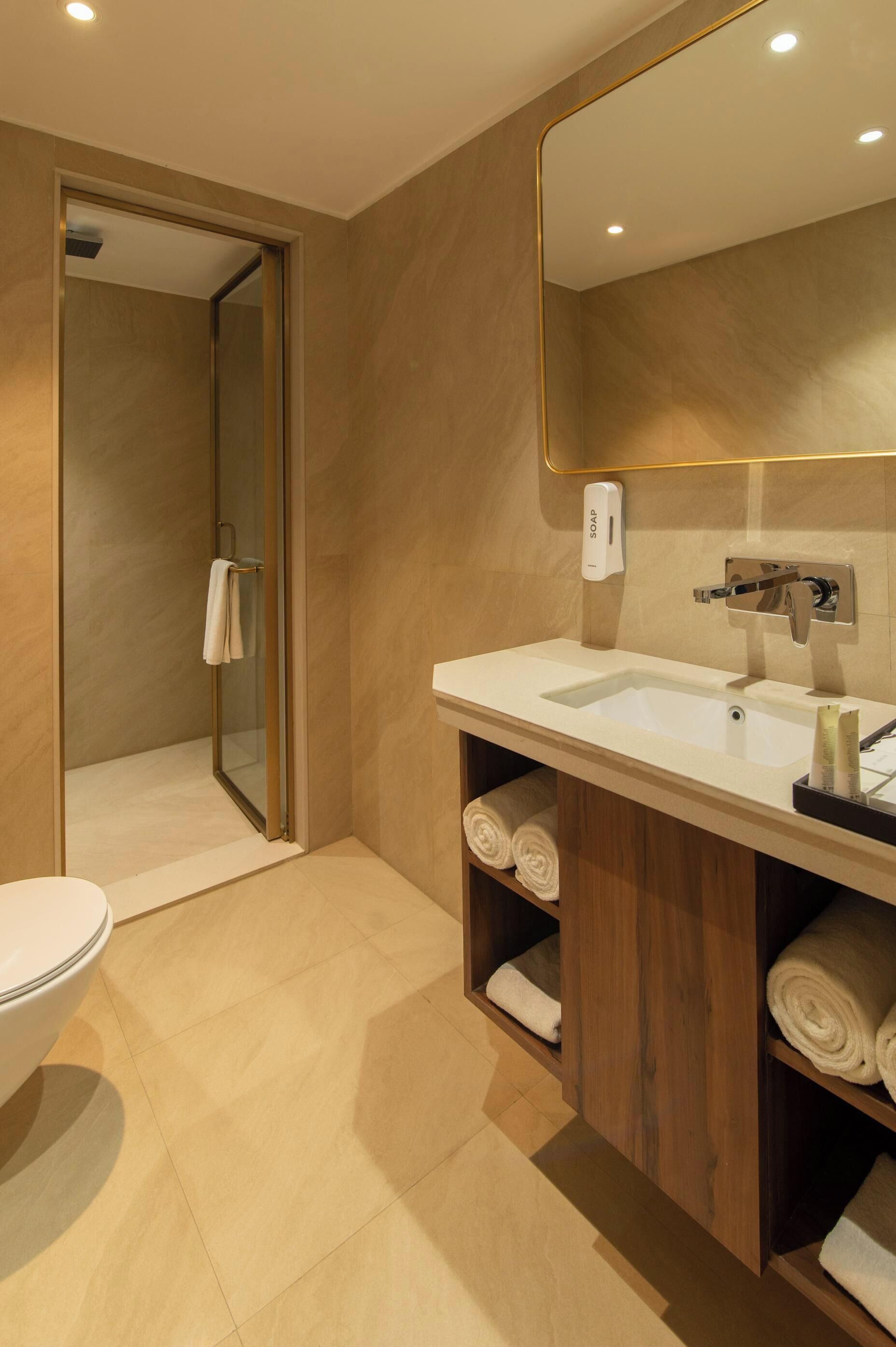 A modern bathroom with a white sink, a mirror, and a shower. There are towels neatly rolled on shelves.
