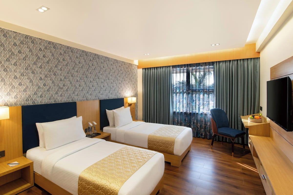 A hotel room with two beds, a desk with a chair, a TV, and a window with curtains.