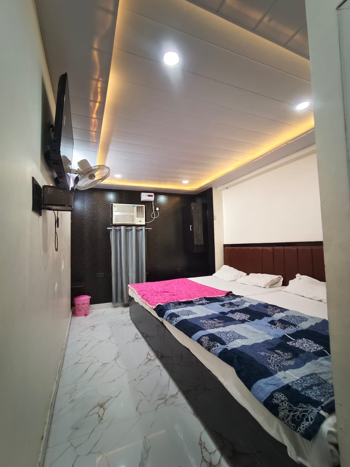 Quarto de hotel com cama, TV fixada na parede, ar condicionado e ventoinha no teto.