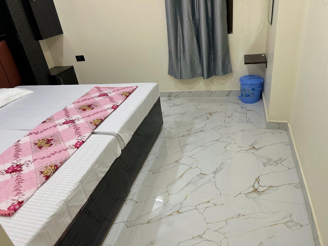 A hotel room with a bed, a marble floor, a blue bucket, and a grey curtain.