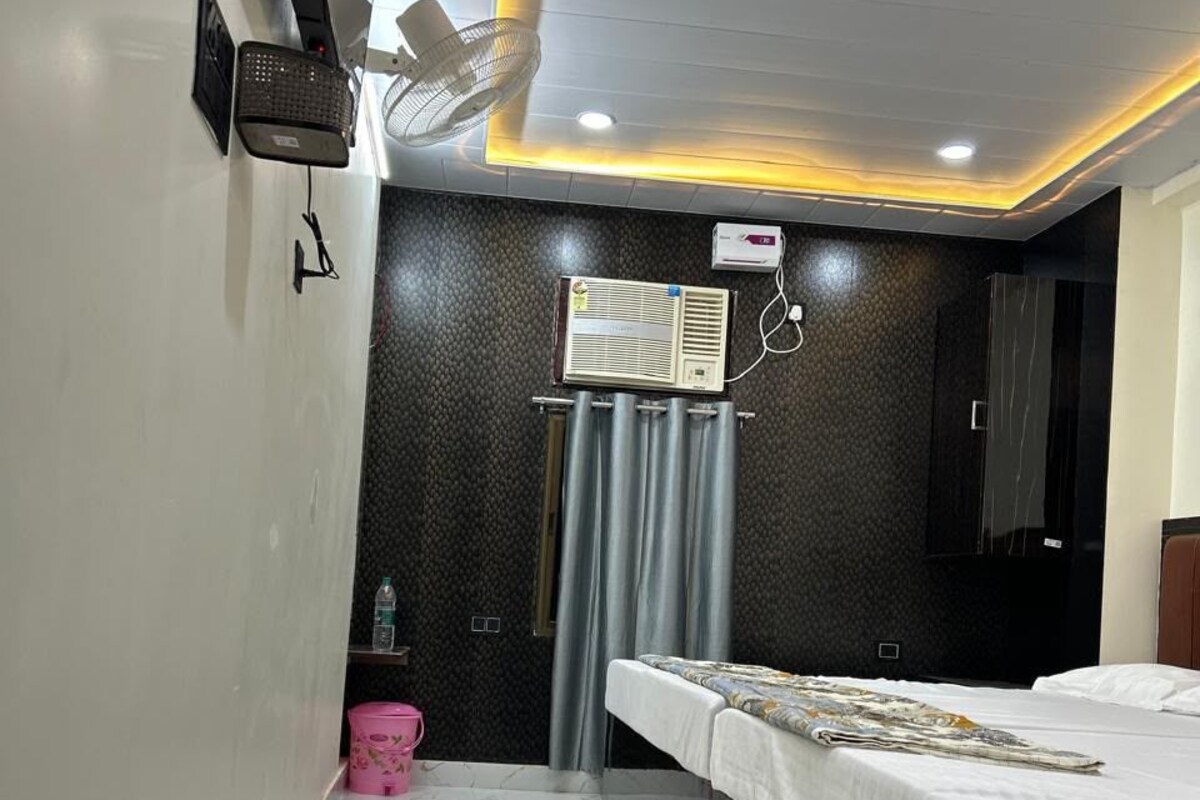 A hotel room with a bed, a ceiling fan, a wall-mounted air conditioner, and a small shelf with a water bottle.