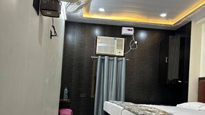 A hotel room with a bed, a ceiling fan, a wall-mounted air conditioner, and a small shelf with a water bottle. - New Jay Radhe Krishan Residency (Varanasi)
