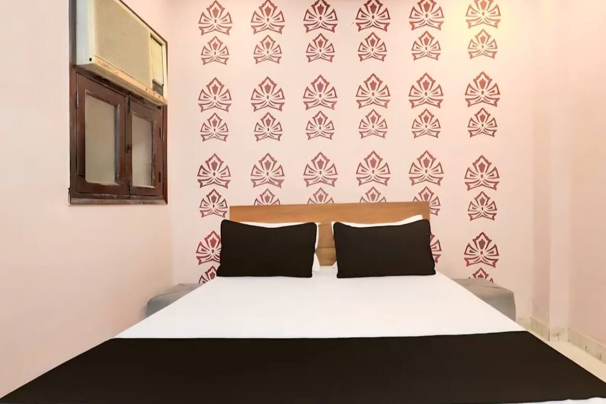 A bedroom with a bed, two black pillows, a wooden headboard, a window with blinds, and a patterned wallpaper.