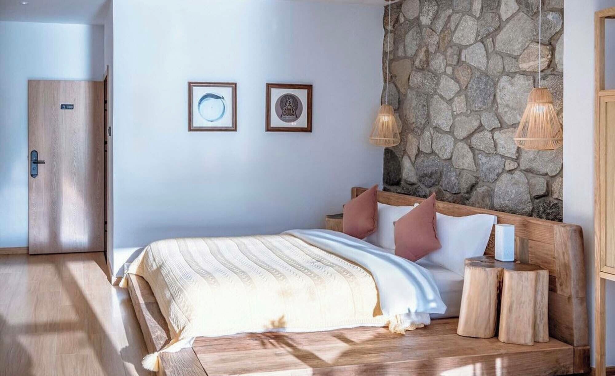 A bedroom with a stone wall, a bed with a wooden headboard, two framed pictures on the wall, and a wooden wardrobe.