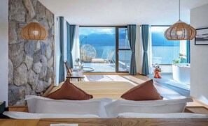 A modern hotel room with a stone wall, a large bed with brown pillows, a dining area with a table and chairs, and a view of the sea through large windows. - Dali Nanshenan Seaview Resort Villa (Dali)