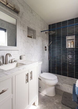 Un baño moderno con tocador blanco, un espejo grande, inodoro y zona de ducha con azulejos negros.
