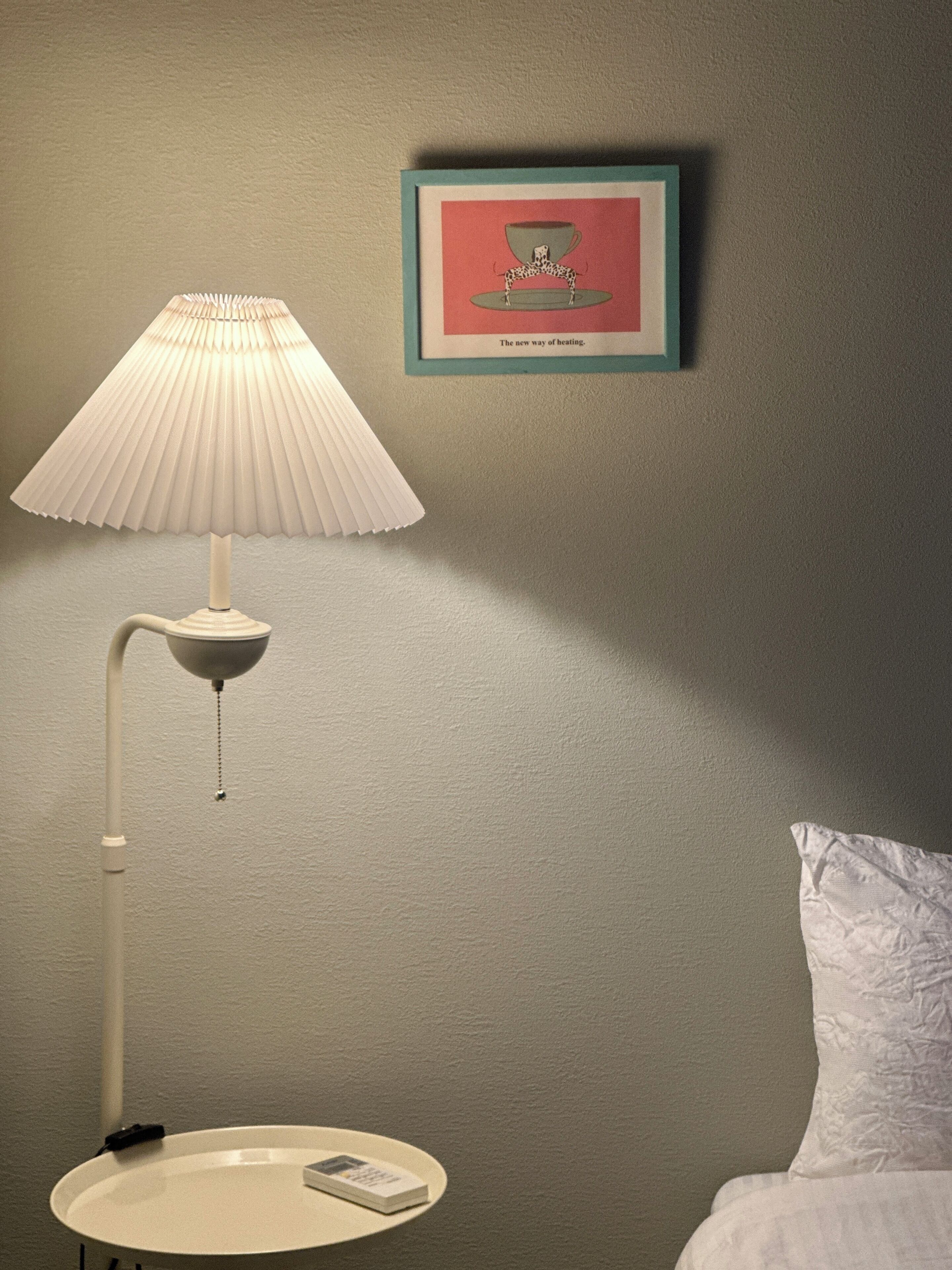 A room with a white bedside table, a white lamp, a framed picture on the wall, and a white pillow.