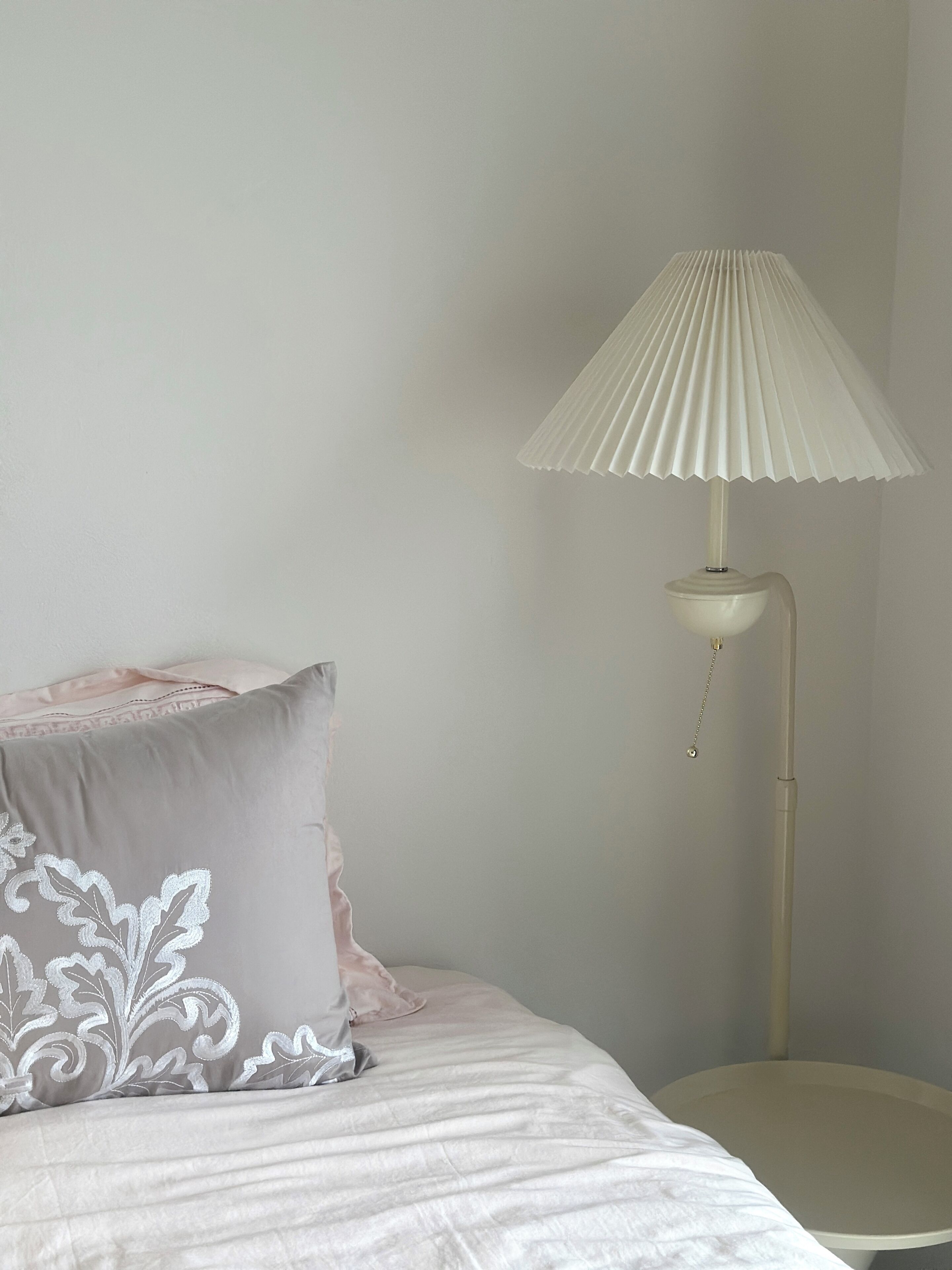 A bed with a white and gray pillow, a white lamp with a pleated shade, and a plain wall.