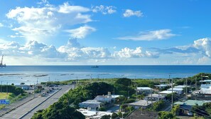 A coastal area with a road, parked cars, and a ship in the distance. - SunSea Okinawa Stunning Ocean View 2BR Sleeps 6 20 min from Airport 3 min to Shopping Mall (Urasoe)