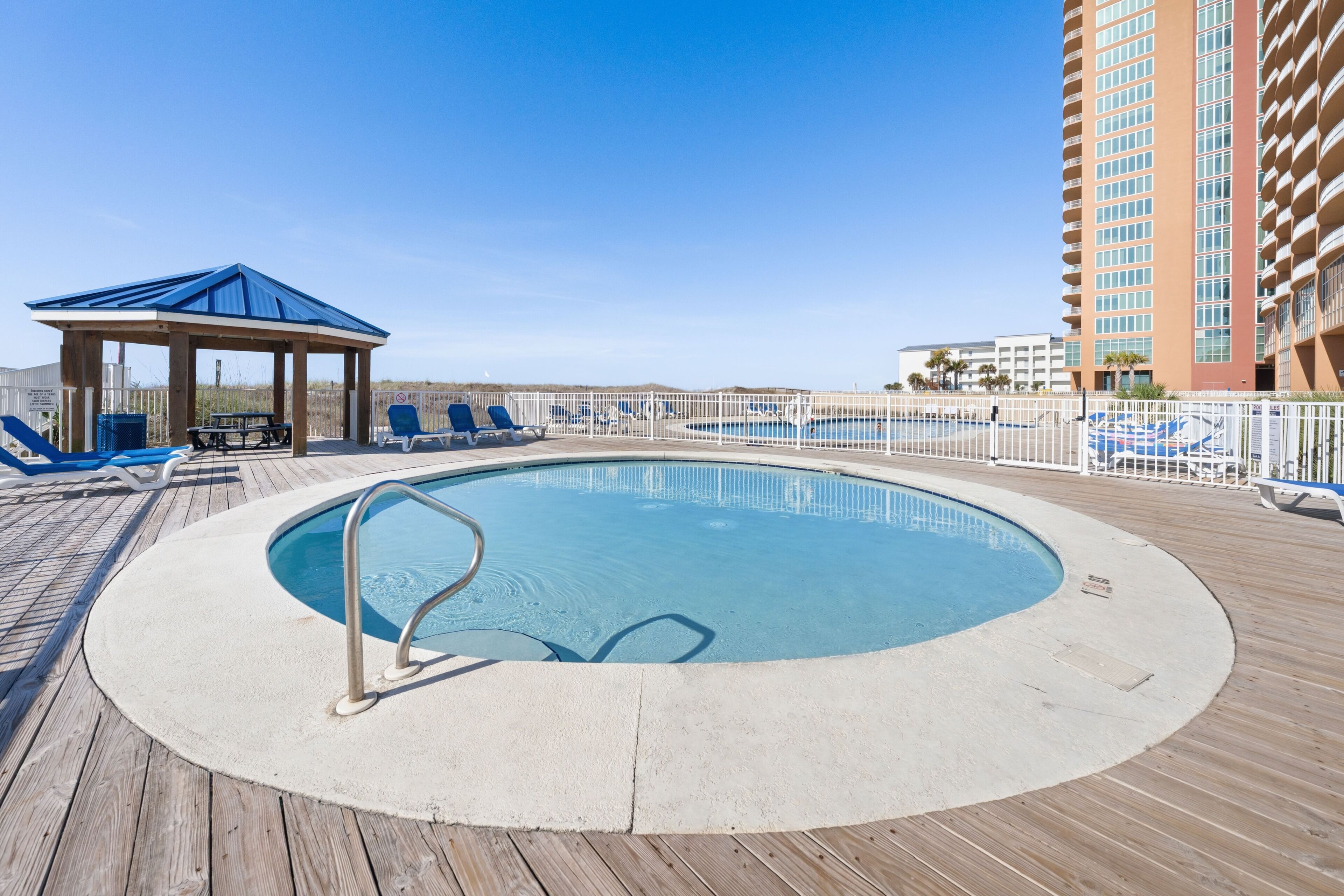A pool area with a wooden deck, lounge chairs, a covered seating area, and a tall building in the background.