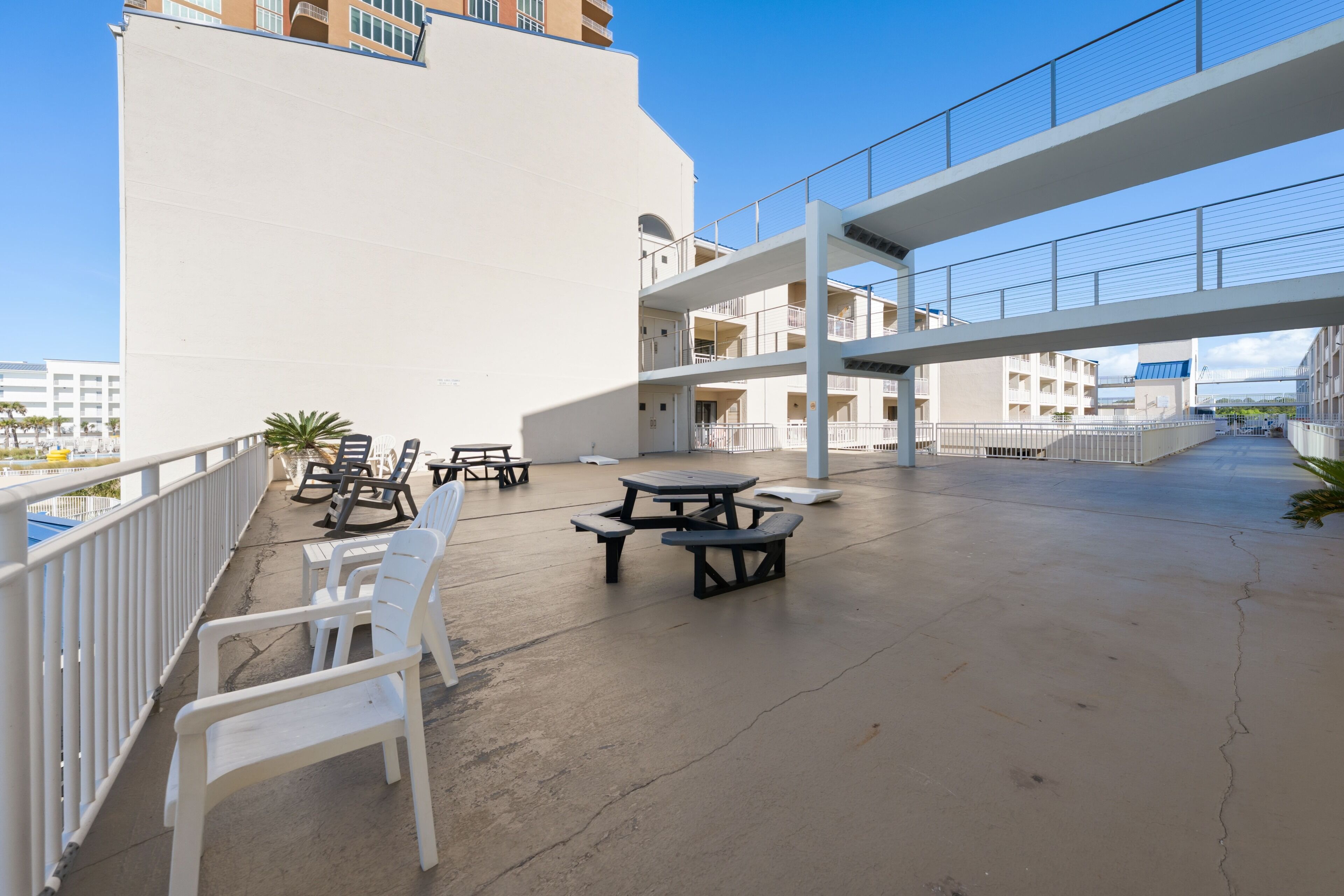 An outdoor patio area with white plastic chairs and tables, a white building with balconies, and a clear blue sky.