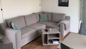 Ein modernes Wohnzimmer mit einem grauen Ecksofa, einem kleinen weißen Couchtisch und einem Essbereich mit Tisch und grauen Stühlen.