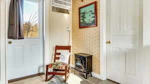 Een kamer met een witte deur, een stoel met een bloemenkussen, een kleine houtkachel en een wandairconditioner.