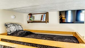 Een kleine, knusse kamer met een enkel bed, een boekenkast, een wandtelevisie en een rookmelder aan de muur.