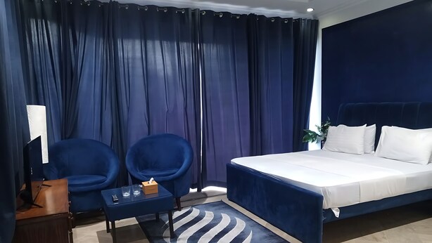 A hotel room with a bed, two blue armchairs, a small table with a tissue box, a lamp, and a large window with curtains. - Sapphire Lodge (Islamabad)