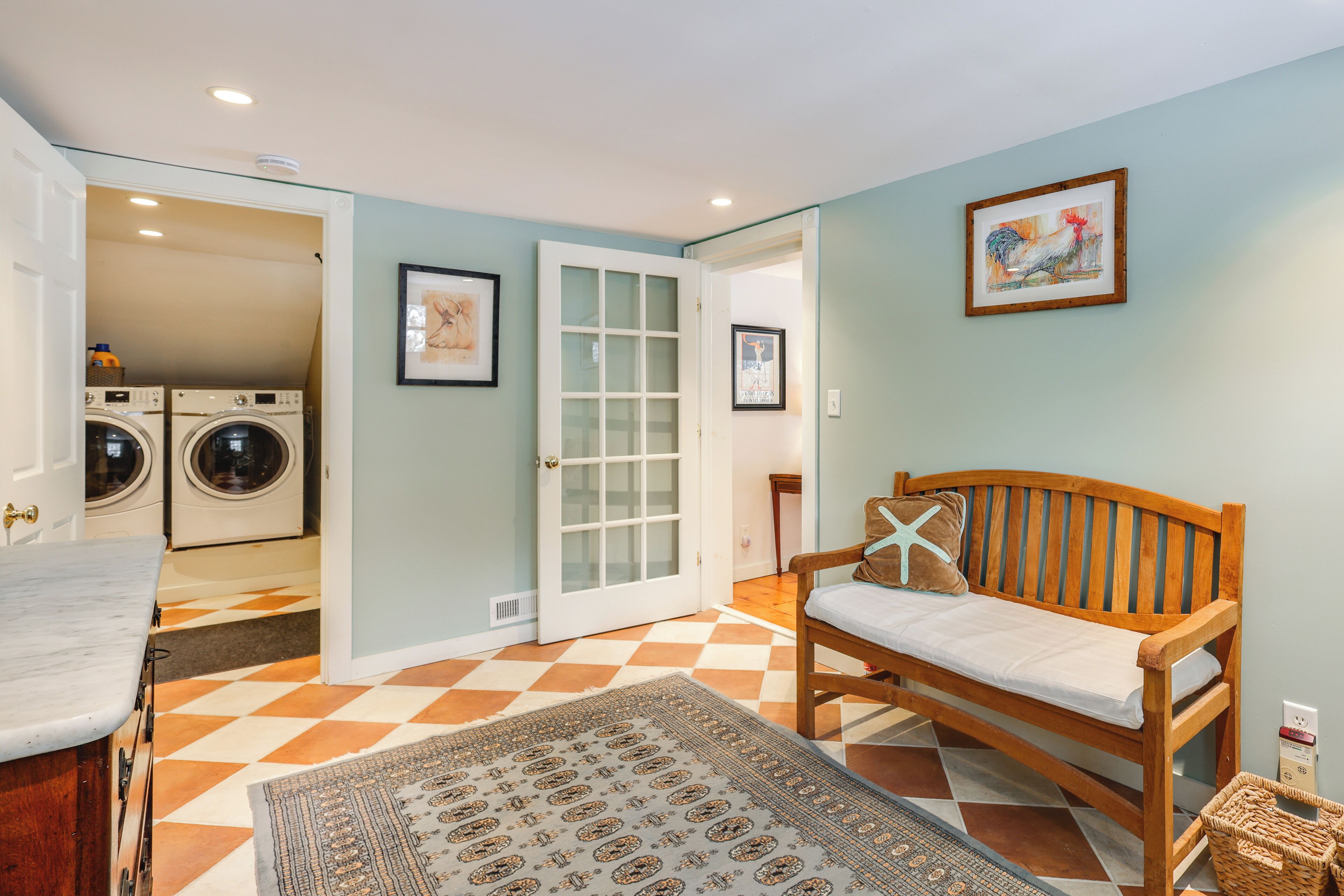 A laundry room with a washing machine and dryer, a wooden bench with a cushion, and a patterned rug on a checkered floor.