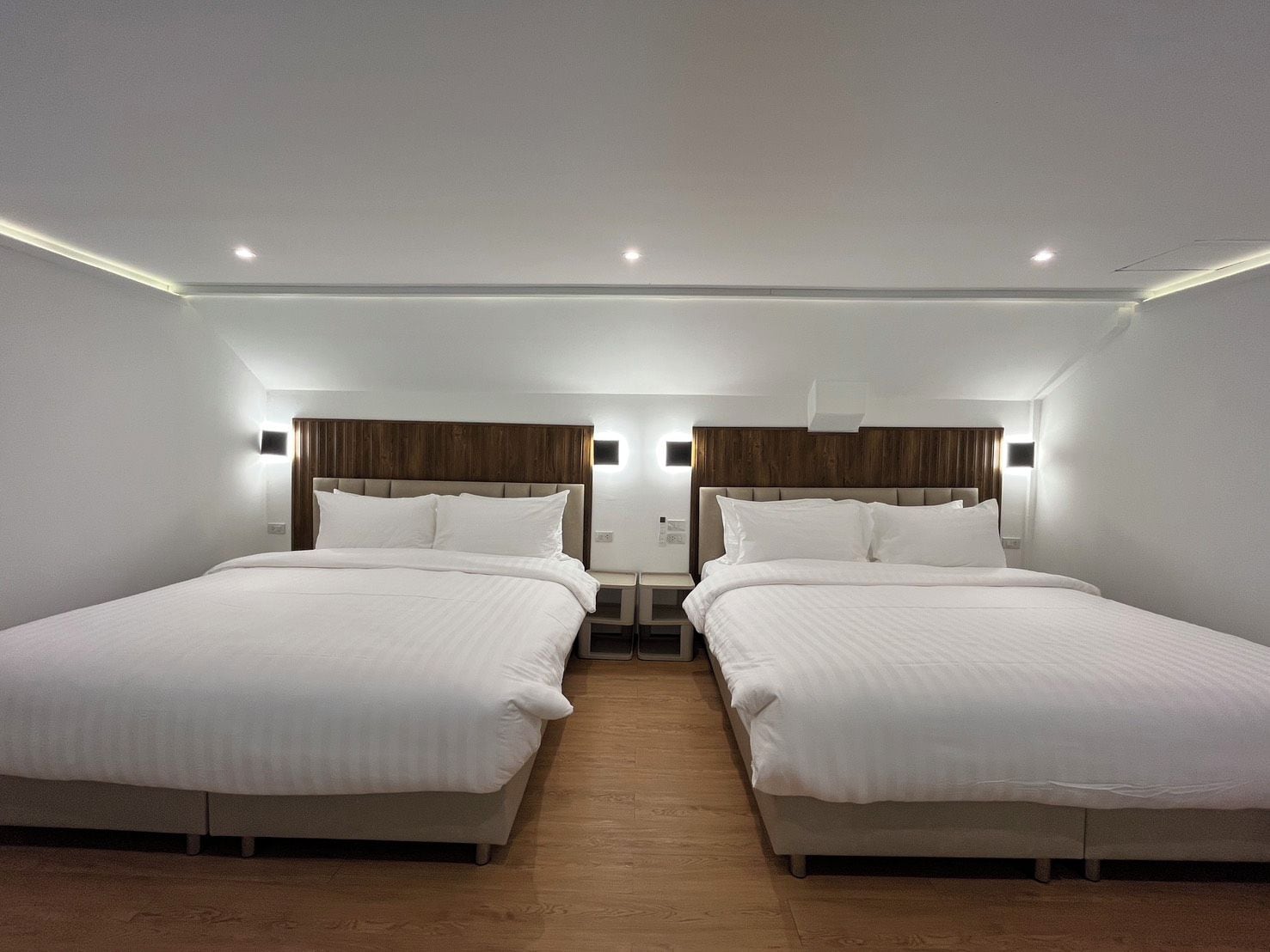 A hotel room with two beds, wooden flooring, and modern lighting fixtures.