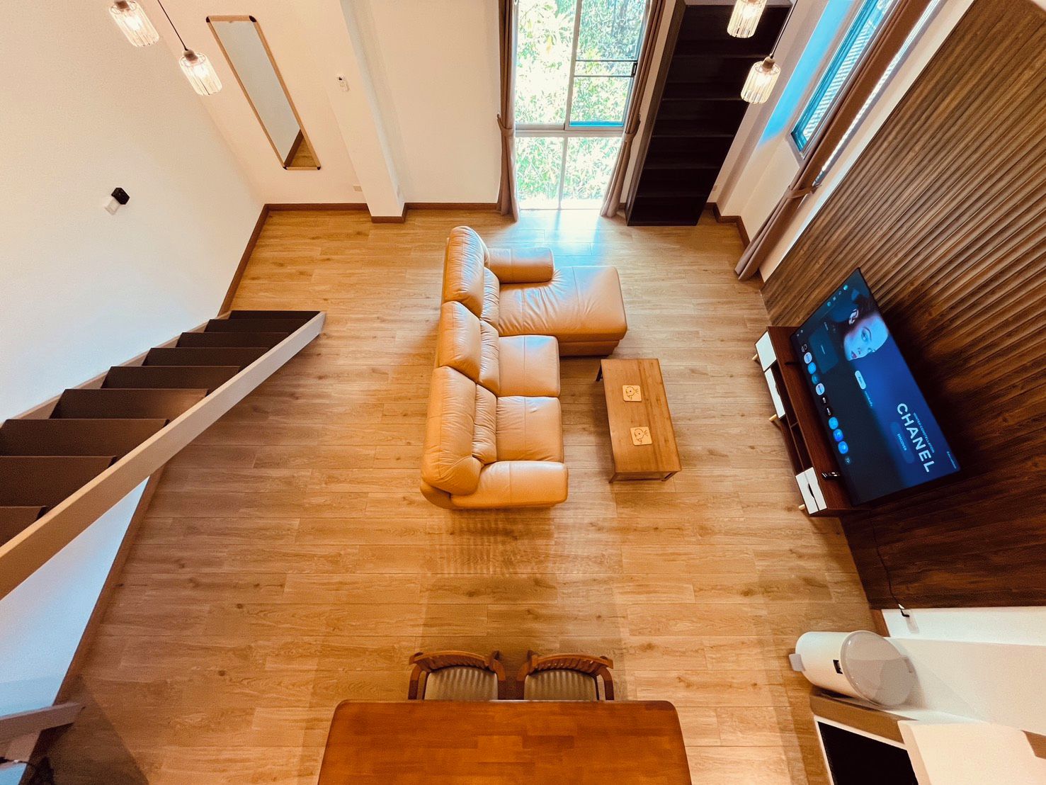 A spacious living area with a leather sofa, wooden coffee table, and a TV displaying a Chanel advertisement. A staircase with wooden steps is visible on the left.