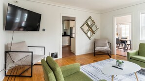 A modern living room with a flat-screen TV, green and white chairs, a round coffee table, and a kitchen visible through an open doorway. - Quiet Room + Great Location Near UW (Seattle)