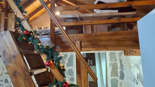 Una escalera de madera adornada con luces y adornos, que conduce a un área tipo altillo con vigas a la vista y una pared de piedra.