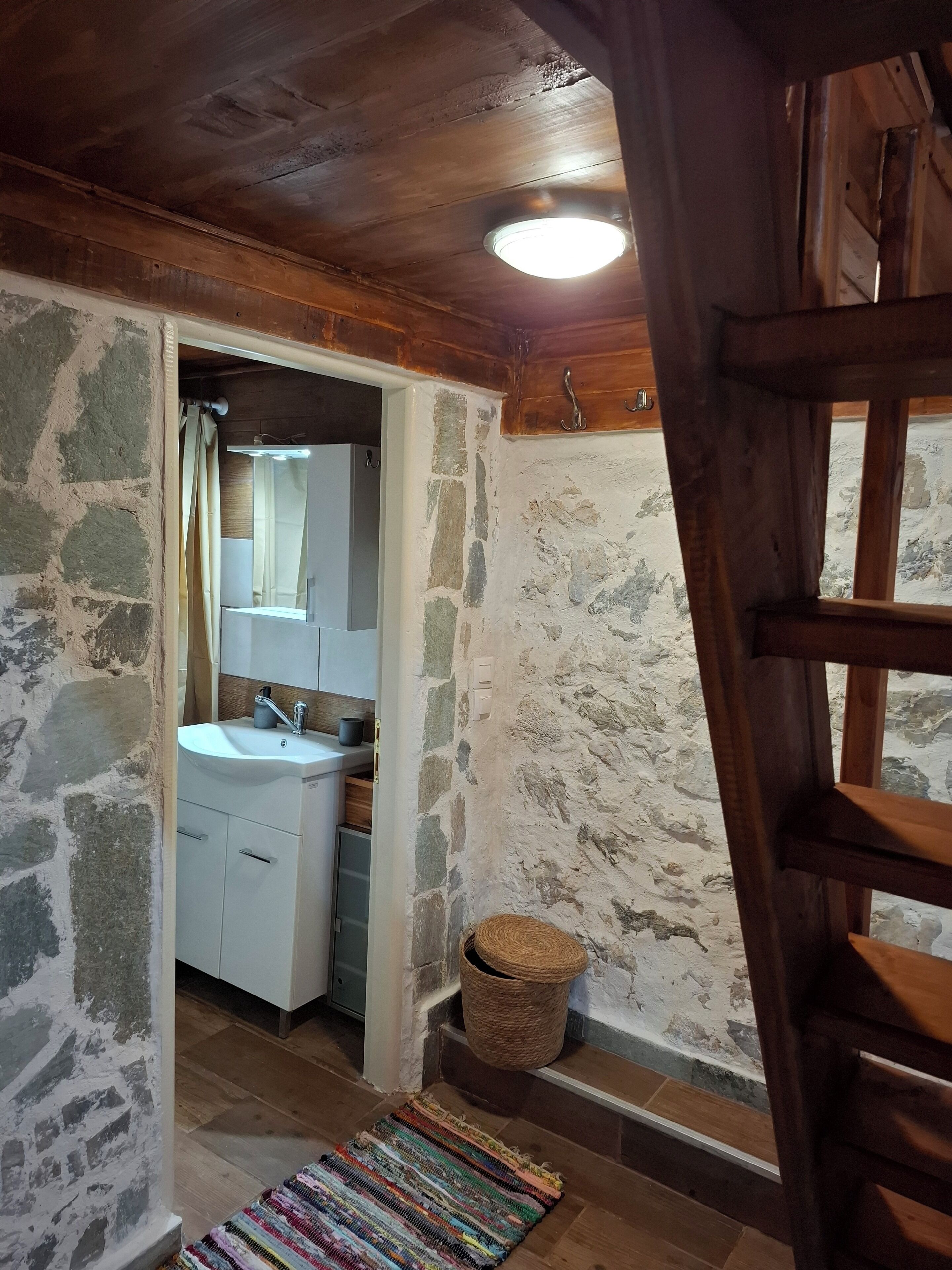 Un baño rústico con pared de piedra, techo de madera y un lavabo blanco con espejo arriba.