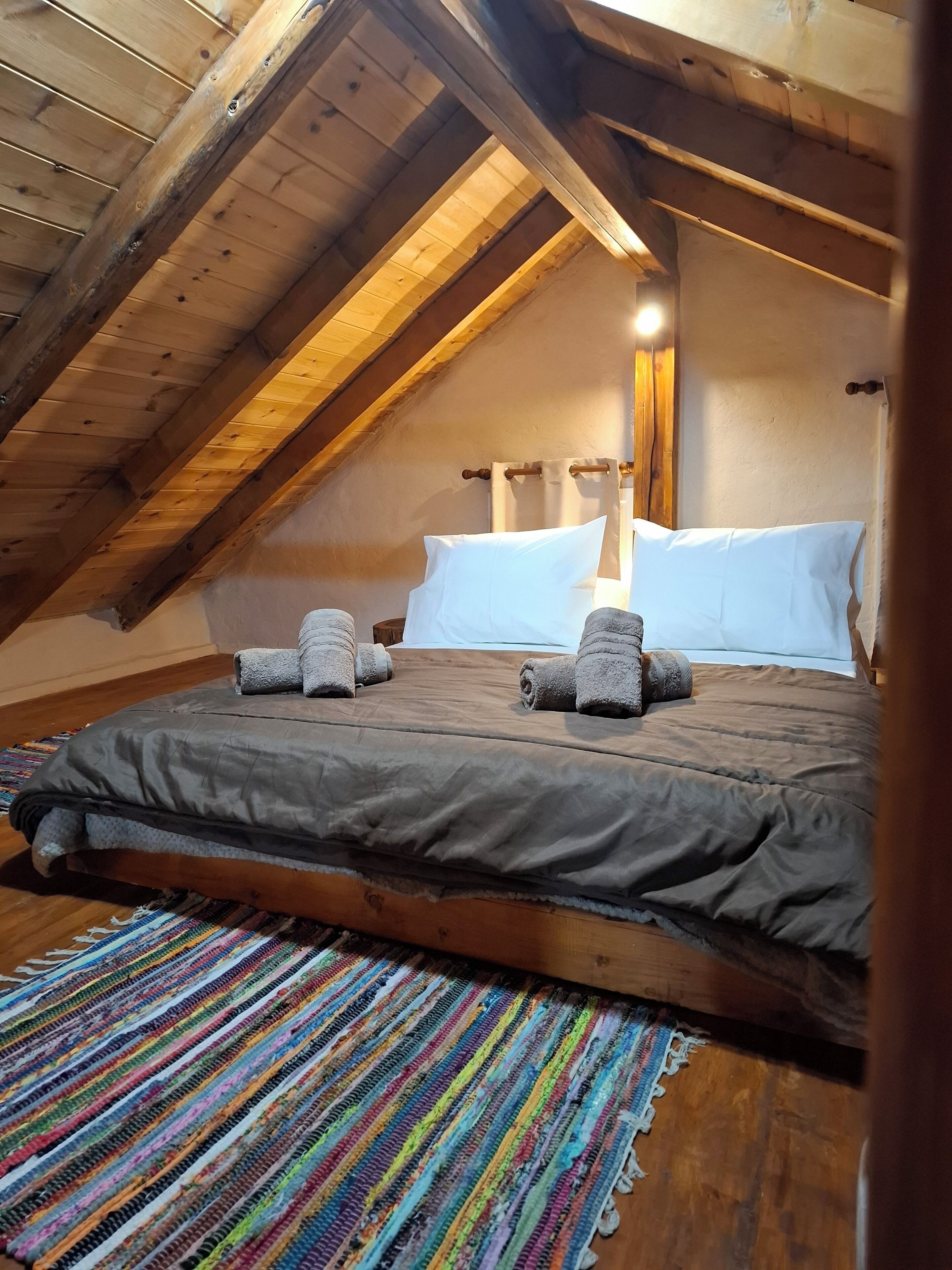 Et hyggeligt soveværelse i en stueetage med skråvægge, træbjælker, en stor seng med gråt sengetøj og en farverig vævet måtte.