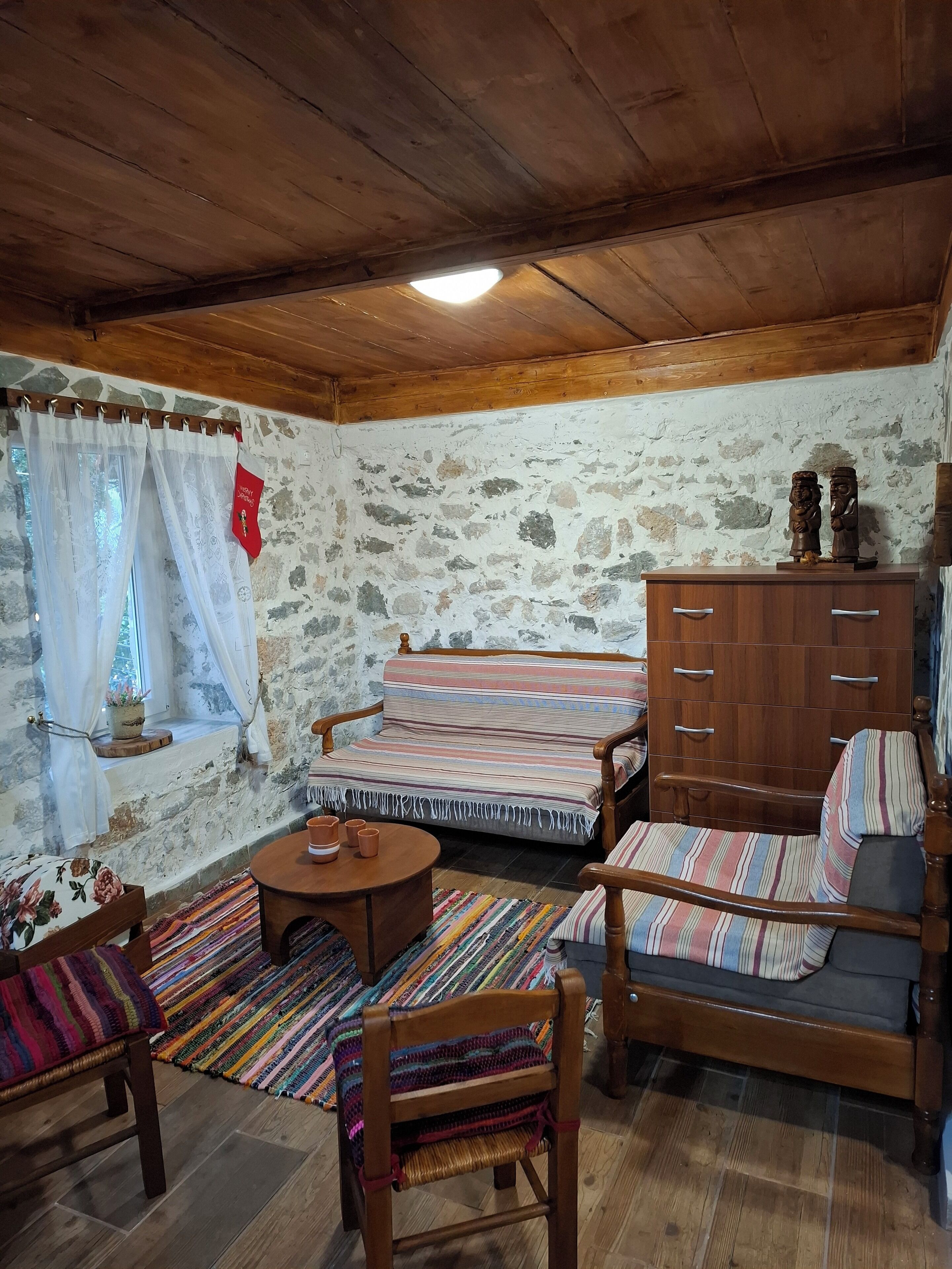 A cozy room with a wooden ceiling, a stone wall, a sofa, a wooden chair, a small table, and a cabinet.