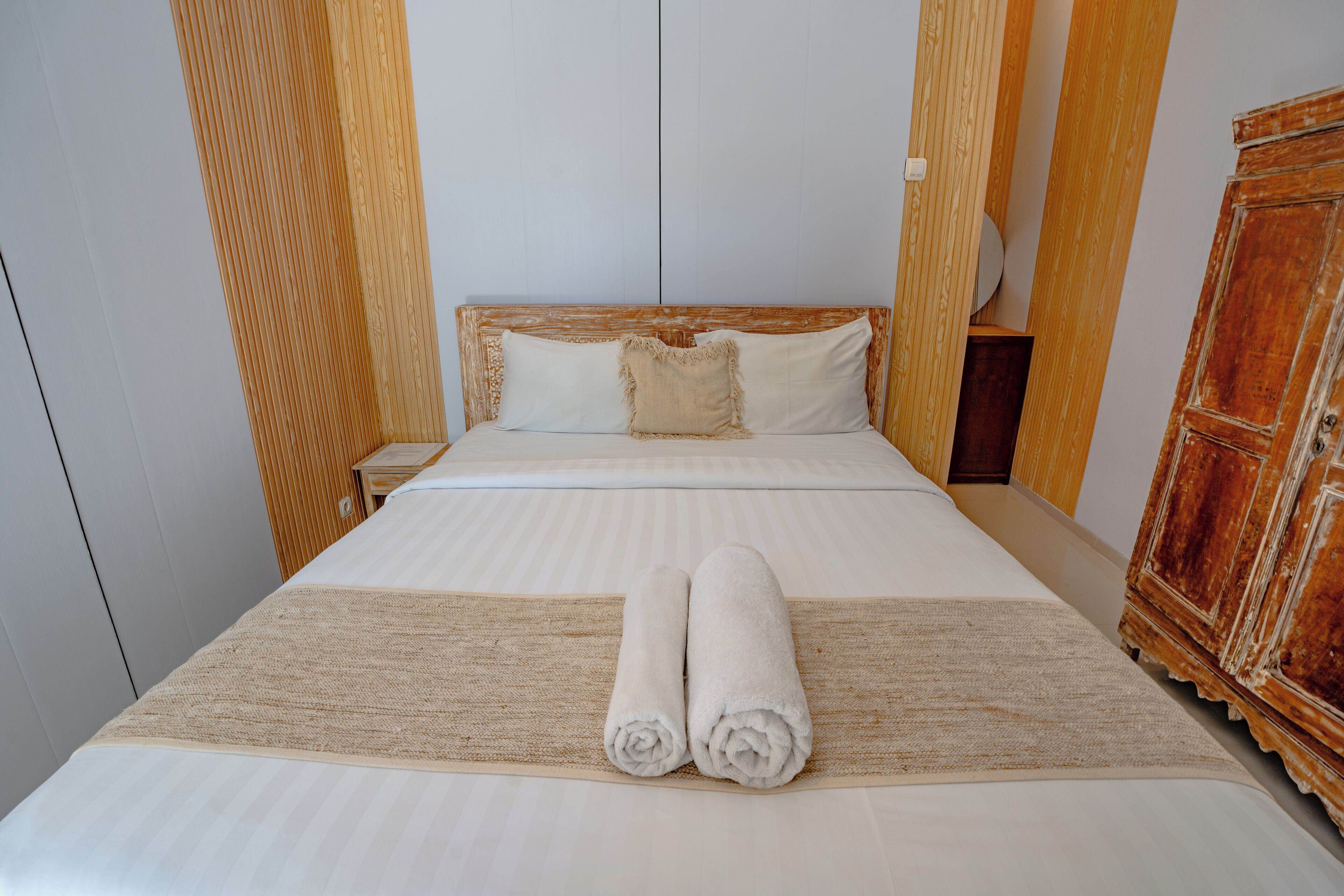 A neatly made bed with two rolled towels, a wooden headboard, a wooden wardrobe, and a plain wall.