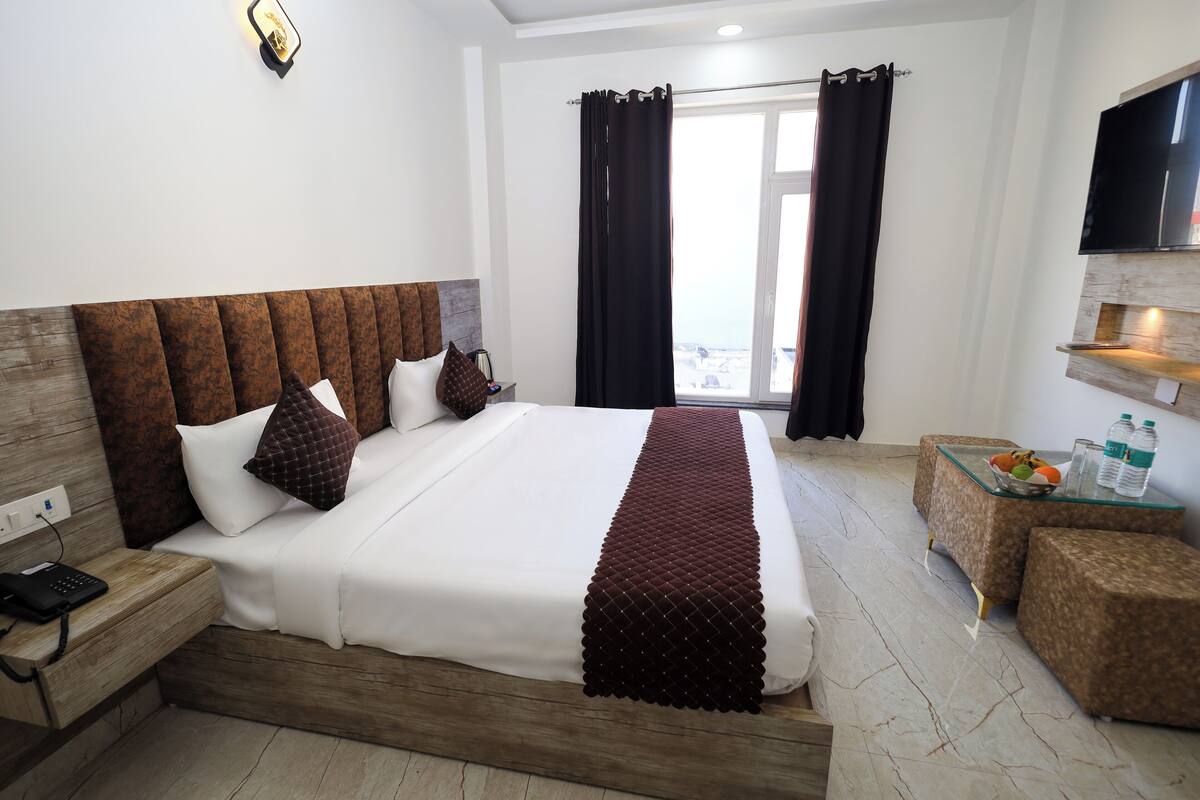 A hotel room with a large bed, a bedside table with a phone, a small table with fruit and bottles, and a TV on the wall.