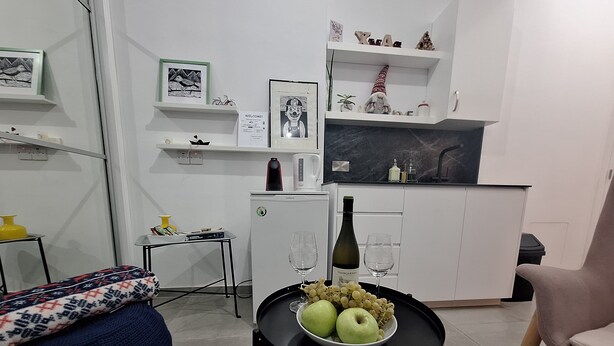 A modern living room with a sofa, a small table with a bowl of fruit and wine, a kitchen area with a fridge, and a dining table. - The studio offers cleanliness, peace and comfort, for a pleasant, relaxing stay! (Pafos)
