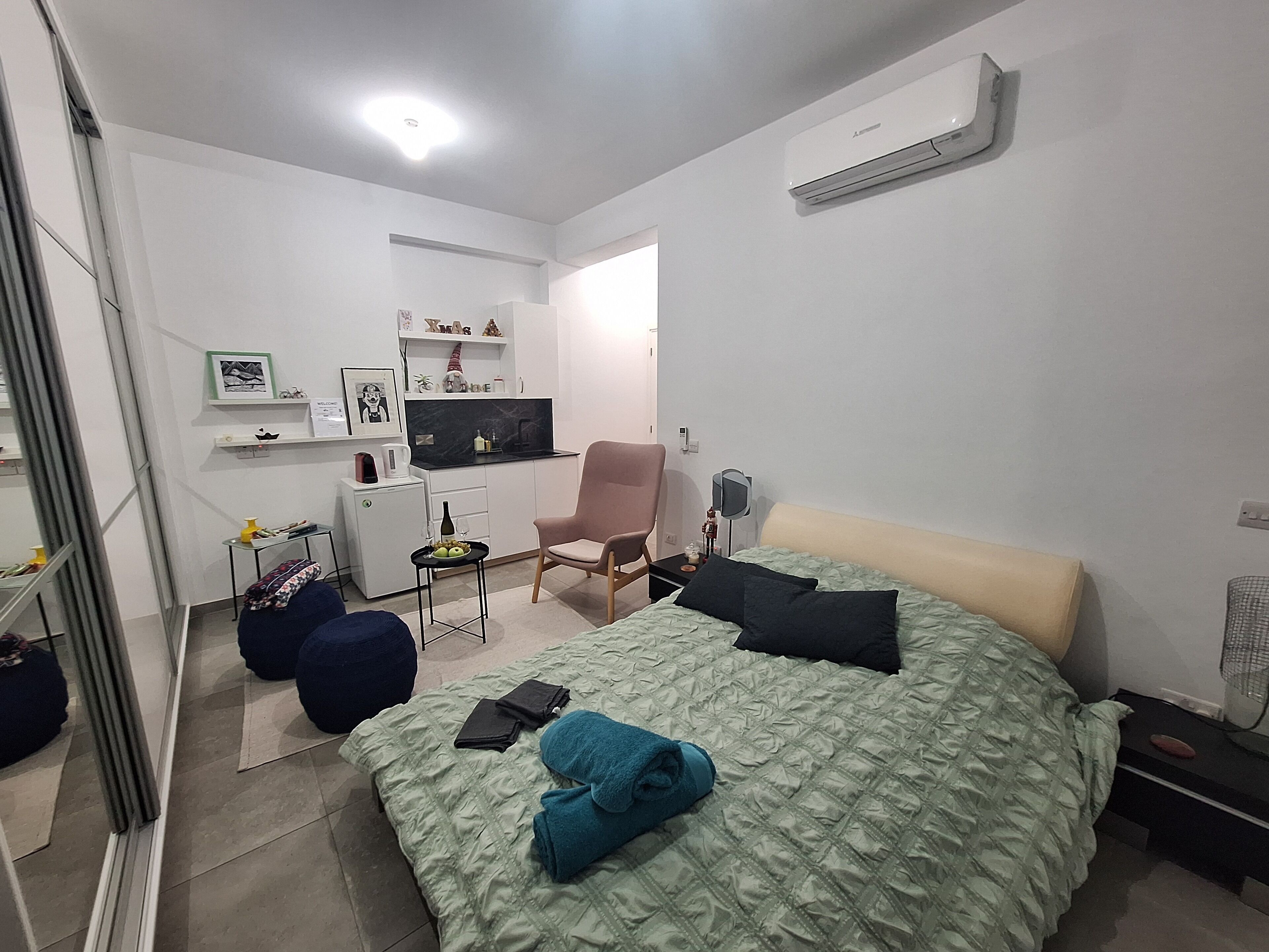 A compact bedroom with a bed, a chair, a small kitchenette, and a mirror.