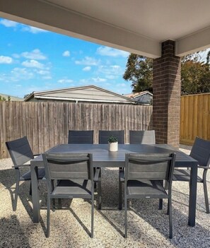 Eine überdachte Terrasse mit einem rechteckigen Tisch und sechs Stühlen, einer Ziegelstütze und einem Holenzaun im Hintergrund.
