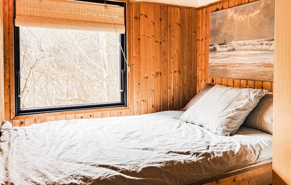 A wooden room with a bed, a window with a view of trees, and a painting of a beach scene.