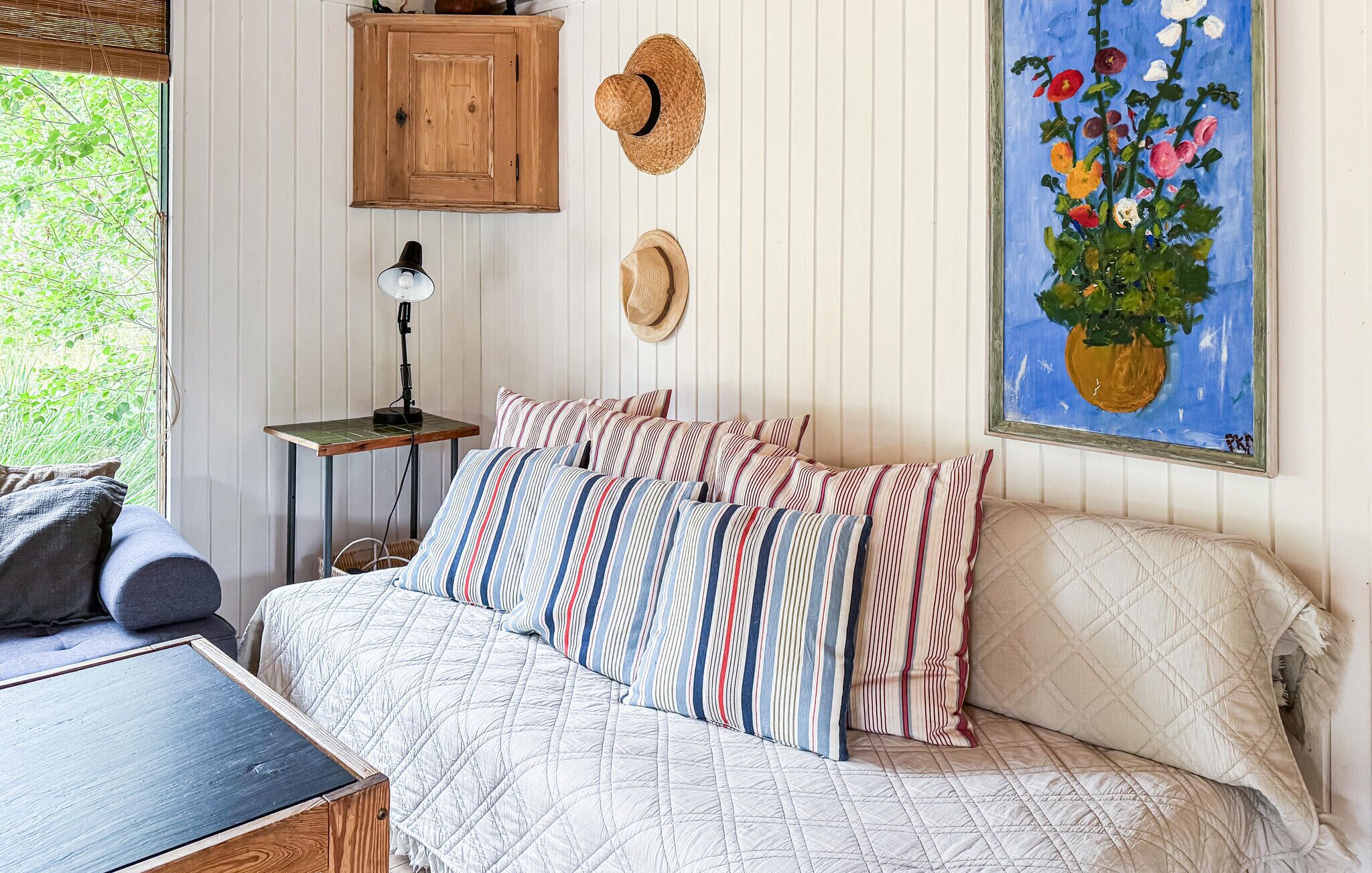 A bedroom with a bed, pillows, a nightstand, a lamp, a hat, and a painting.