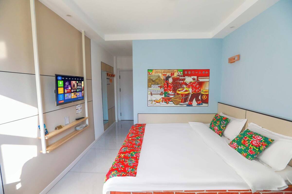 Een hotelkamer met een groot bed, een aan de muur bevestigde televisie, een schilderij van een traditioneel tafereel en een gang naar een andere kamer.