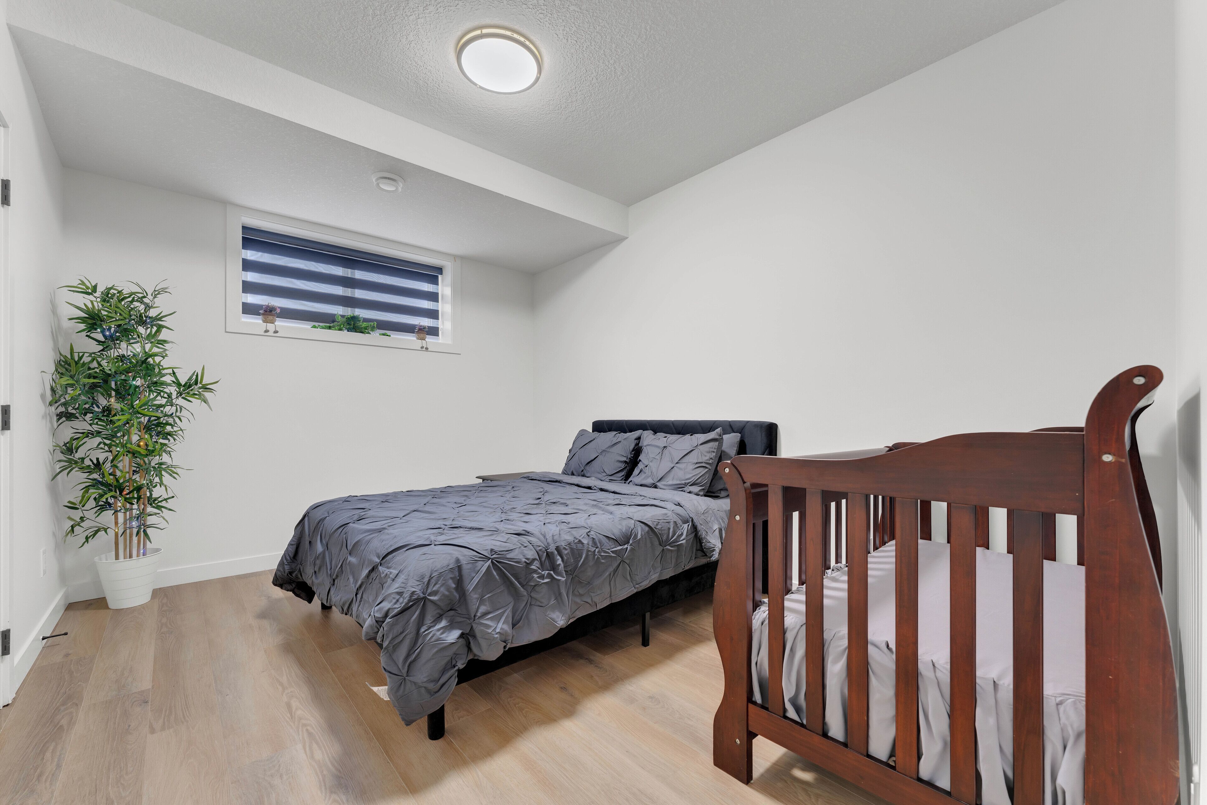 Ein Schlafzimmer mit einem Bett, einem Babybett, einer Zimmerpflanze und einem Fenster mit Jalousien.
