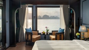A hotel room with a large window, two chairs, a small table with fruit, and a bed. - Aime'e Luxury Cruise (Bai Chay)