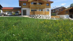 A two-story wooden house with a balcony, a stone foundation, and a clear view of the mountains in the background.