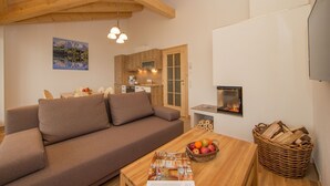 A cozy living room with a sofa, a wooden coffee table with fruit, a fireplace, and a kitchen in the background.