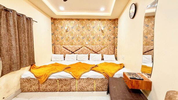 A bedroom with a patterned headboard, a bed with white linens and a yellow throw, a wooden nightstand, and a mirror on the wall. - Premium Hotel Sipra near Mahakal Temple  (Ujjain)