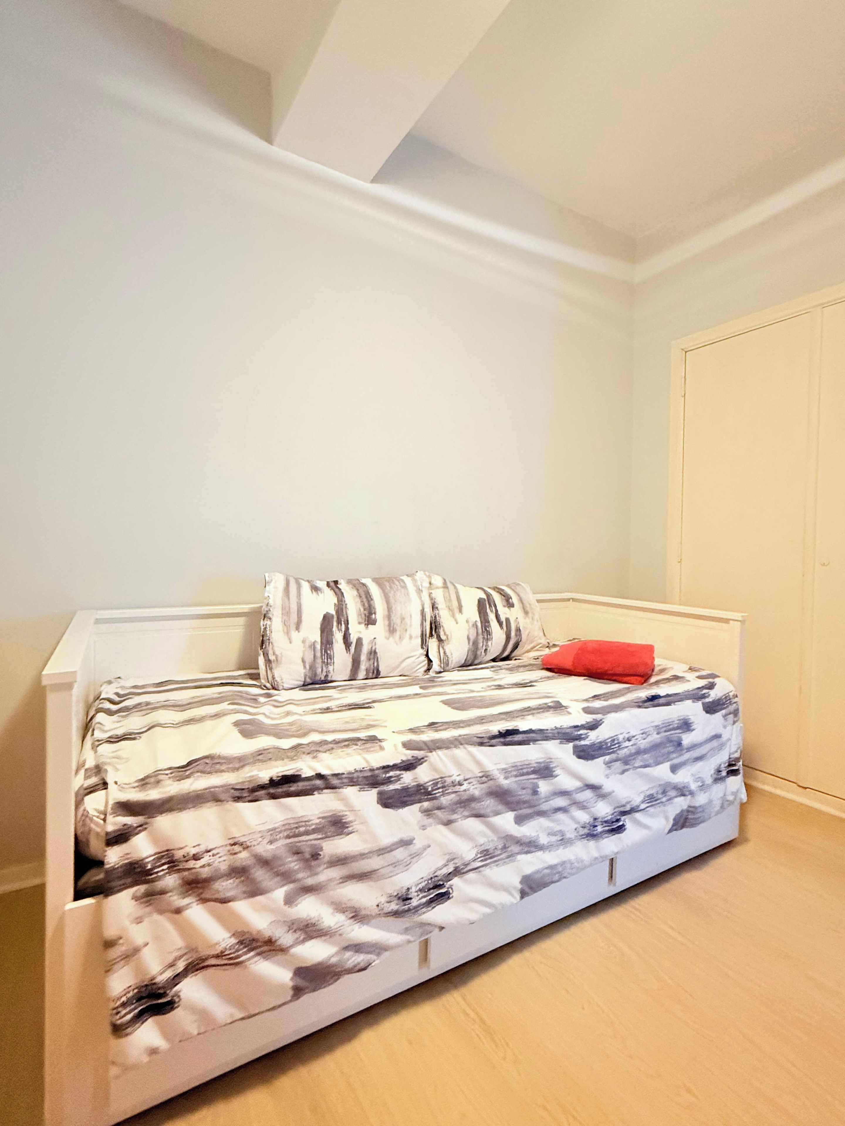 Una cama individual con una colcha estampada, un almohánero rojo y un cabecero blanco en una habitación con paredes blancas y suelo de madera.