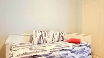 A single bed with a patterned bedspread, a red pillow, and a white headboard in a room with white walls and a wooden floor.